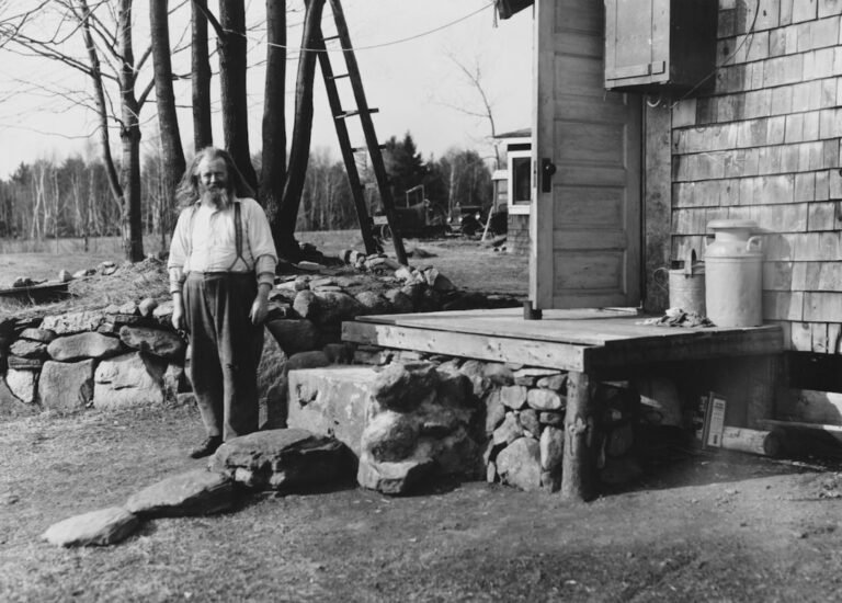 Man standing by a stone wall and wooden building.